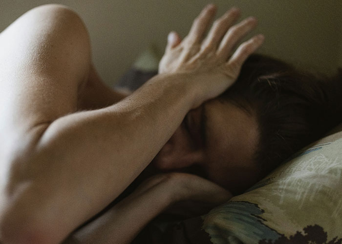 Person covering their face while lying in bed, disturbed by loud music from neighbor late at night.