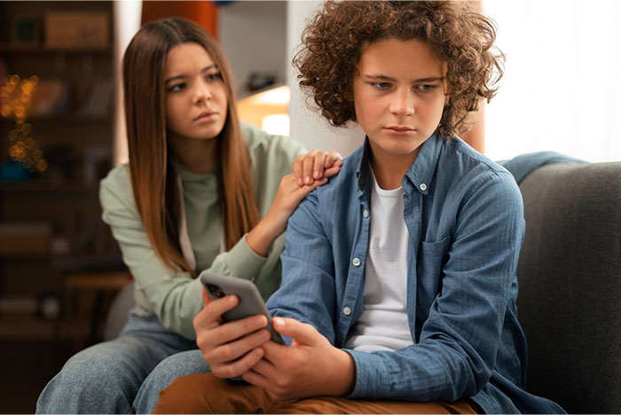 Teen boy looking upset on a couch holding a phone while a concerned woman comforts him at home, highlighting family issues. Teen boy looking upset on a couch holding a phone while a concerned woman comforts him at home, highlighting family issues.