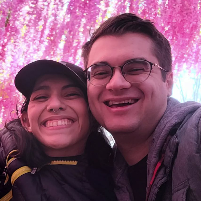Conjoined twin and husband smiling together under vibrant pink flowers, sharing a joyful moment in their marriage.