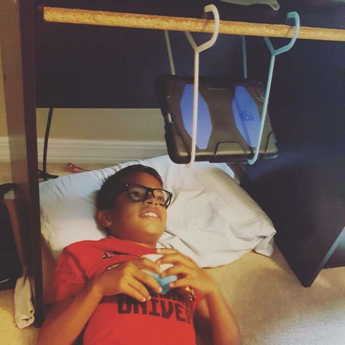 Child with glasses lying under a desk using a homemade tablet holder invention, showcasing kid creativity and innovation.