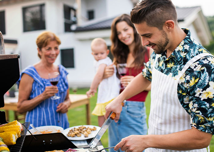 Family barbecue with man grilling food, woman holding toddler, and mother-in-law enjoying wine outdoors in the backyard.