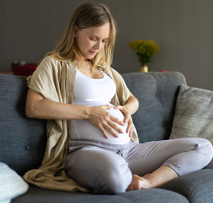 Pregnant woman sitting on couch, gently touching belly, illustrating crazy things heard from MILs needing to calm down.