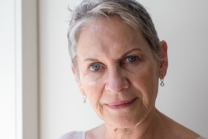 Close-up of a mature woman with short gray hair and blue eyes, expressing calmness amidst crazy MILs comments.
