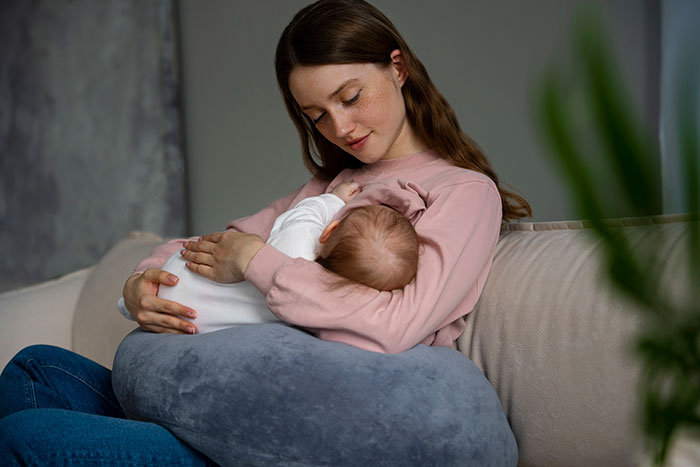 Young mother breastfeeding baby on couch with nursing pillow, illustrating crazy things heard from MILs about parenting.