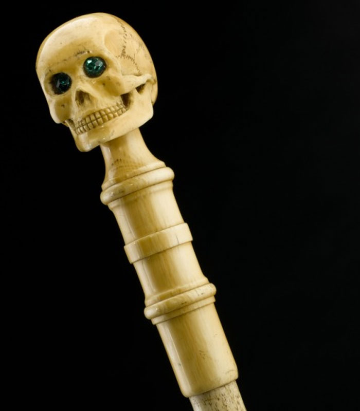 Carved antique ivory staff with skull handle and green gemstone eyes against a black background, history anecdotes artifact.