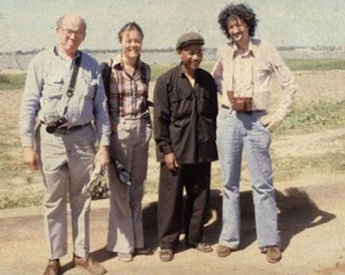 Four people standing outside in historical anecdote setting, smiling and dressed in vintage casual clothing.