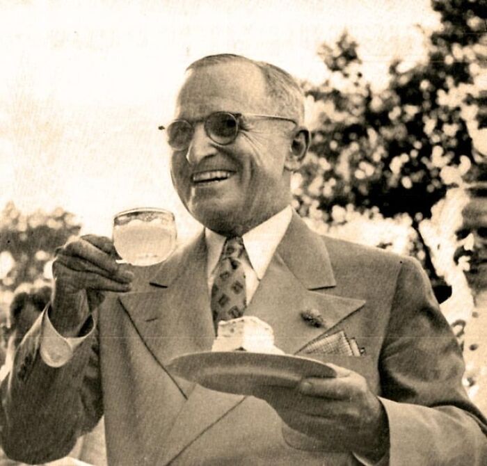 Smiling elderly man in vintage suit holding a drink and plate, captured in a classic history anecdotes moment outdoors.