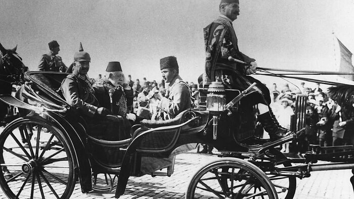 Historical anecdote showing vintage horse-drawn carriage with men in traditional attire and a crowded background.