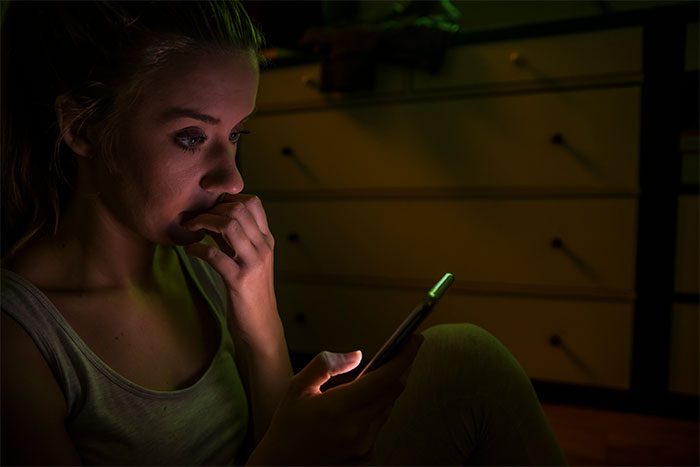 Young woman in dim room looking anxious and staring at phone, experiencing creepy things she can’t explain.