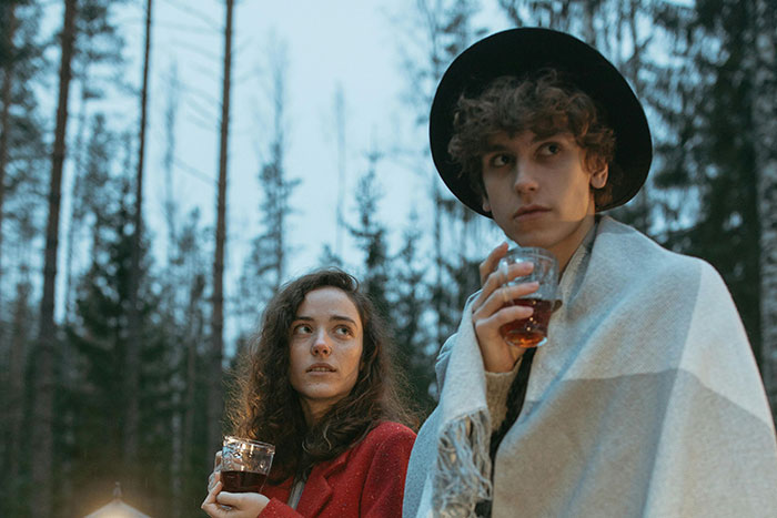 Two people outdoors at dusk holding drinks, surrounded by trees, evoking a creepy and unexplained atmosphere.
