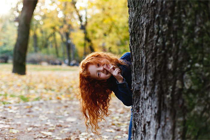 Red-haired girl peeking from behind a tree in a forest during autumn, evoking creepy things people can’t explain experiences.