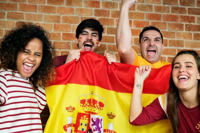 Group of excited young people cheering and holding a Spanish flag, illustrating out of touch moments shared by others.