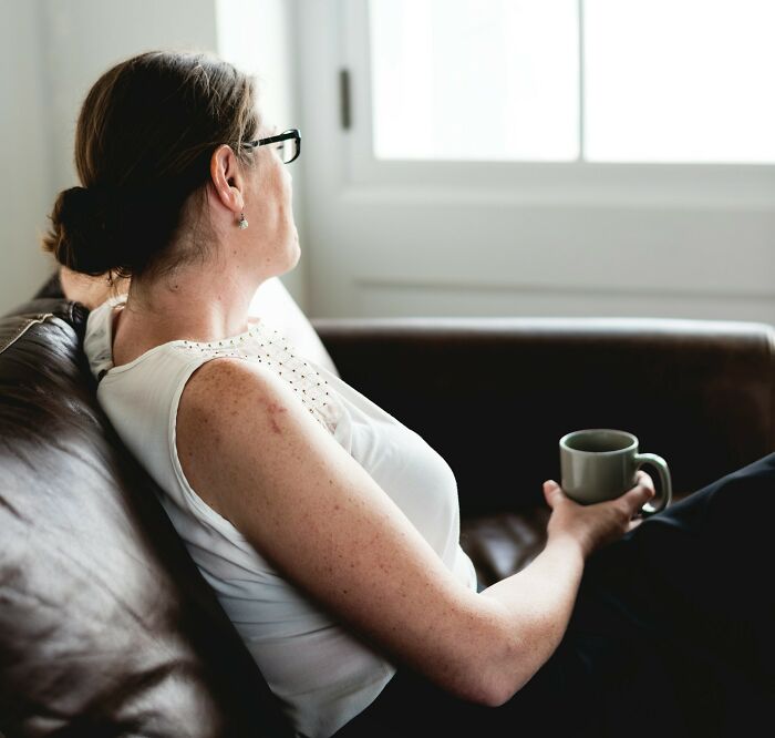 Woman with glasses sitting on a couch holding a cup, reflecting quietly on crazy stories servers overheard.