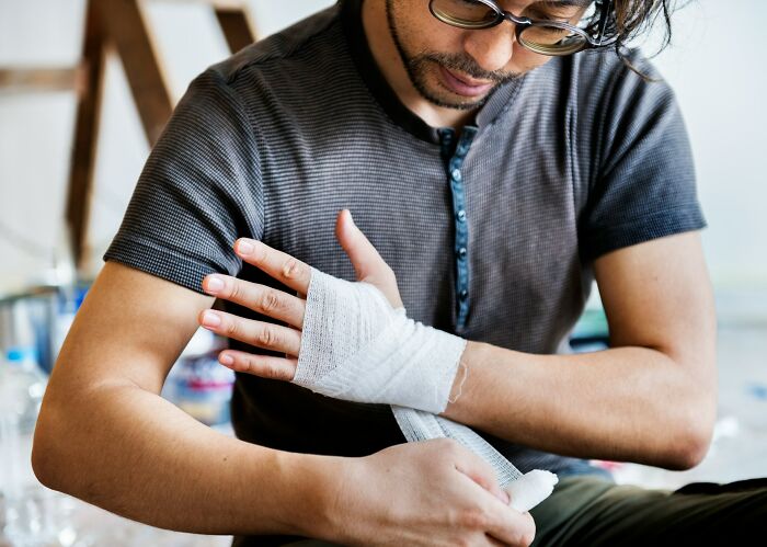 Man with glasses wrapping a bandage around injured hand, illustrating careless mistakes leading to getting fired and blame.