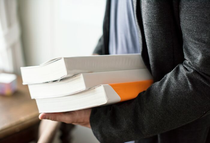 Person in a dark jacket holding several thick books, illustrating insider tricks employees use to make their jobs easier.