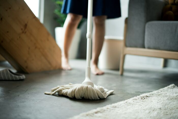 Person using a mop to clean a concrete floor near a rug, showing an everyday thing that humbles fully grown adults.