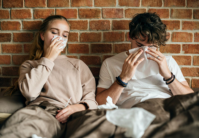 Woman and man sitting on a couch using tissues, appearing unwell due to allergic reaction or anaphylactic shock symptoms.
