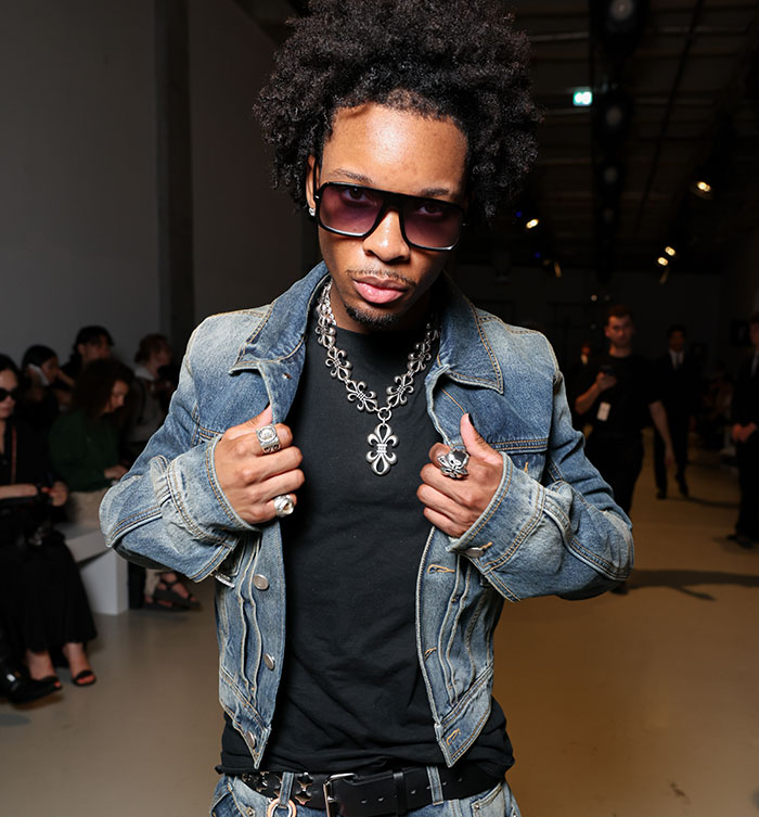 Singer D4vd wearing sunglasses and denim jacket at a public event with people in the background
