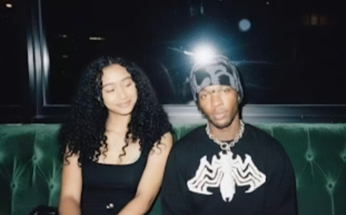 Singer D4vd wearing a beanie and necklace sitting beside a woman with curly hair on a green couch at night.