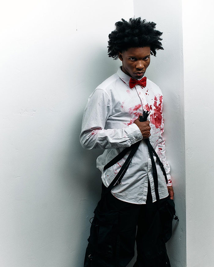 Young man in blood-stained white shirt and red bow tie stands with a serious expression, holding black straps near white wall.