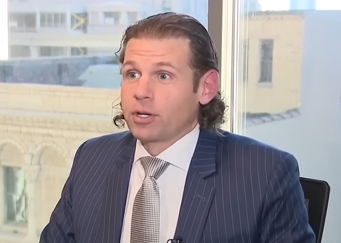 Man in pinstripe suit and patterned tie speaking during an indoor interview about Lyft driver weight discrimination lawsuit.