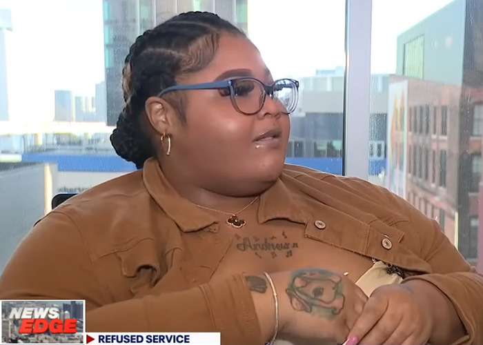 Woman with glasses and brown jacket talks about Lyft driver denying ride due to her weight in an interview setting.