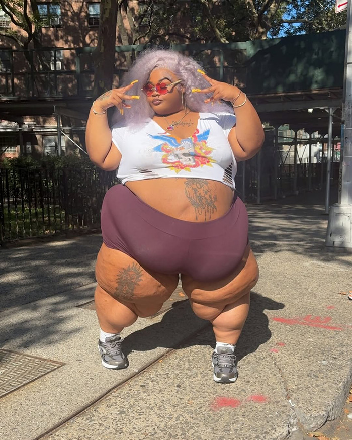 Rapper posing confidently outdoors in casual outfit after Lyft driver denied ride due to her weight settlement news.