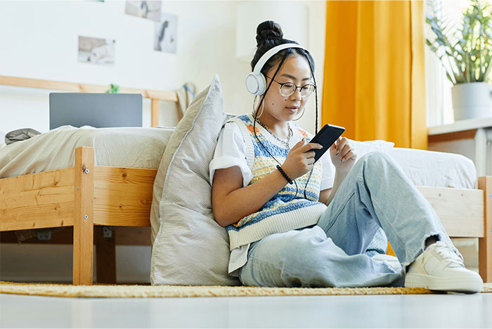 Young girl listening to music with headphones, sitting on the floor in her bedroom with a phone in hand.