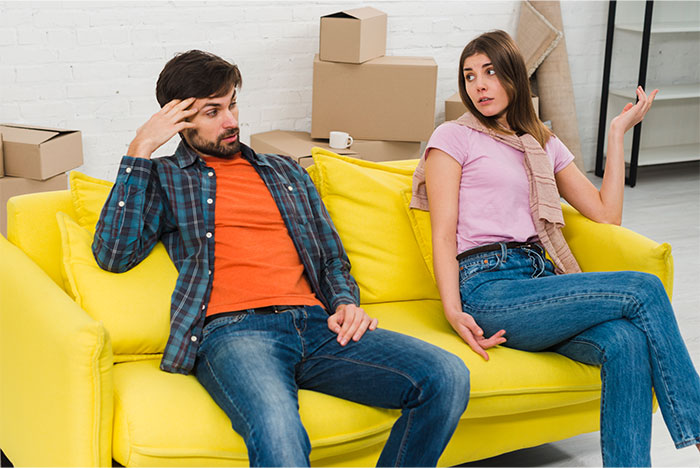 Man and woman sitting on yellow couch in tense conversation, illustrating man defends daughter&rsquo;s room from fianc&eacute;e conflict.