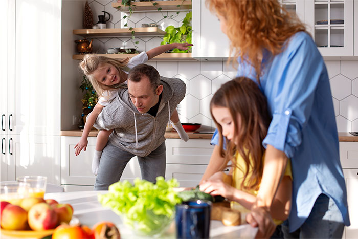 Man playing with daughter in kitchen while another child and woman stand nearby showing family bonding and protection.