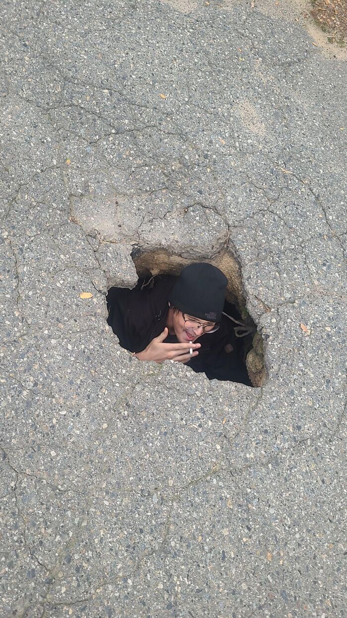 Person wearing glasses and a black hat emerging from a large c*****d pothole, illustrating danger and near-death experience.