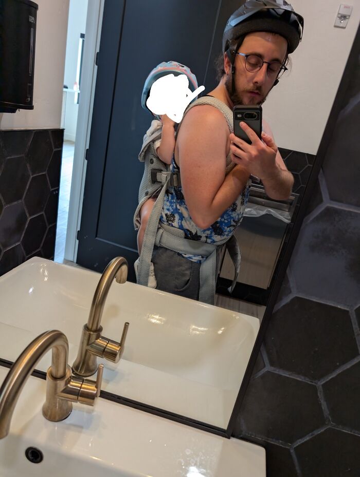 Man wearing helmet carrying child in a baby carrier, taking a mirror selfie in a bathroom with hexagonal tiles.