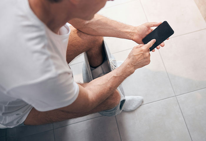 Man sitting on toilet using smartphone in bathroom, illustrating poor hygiene affecting partners and relationships.