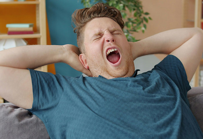 Young man yawning widely on couch, showing tiredness and discomfort related to poor hygiene and gagging reactions.