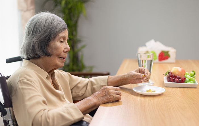 Elderly woman in a beige shirt holding pills at a table with fruit and water, highlighting dementia risk and food concerns. Elderly woman in a beige shirt holding pills at a table with fruit and water, highlighting dementia risk and food concerns.