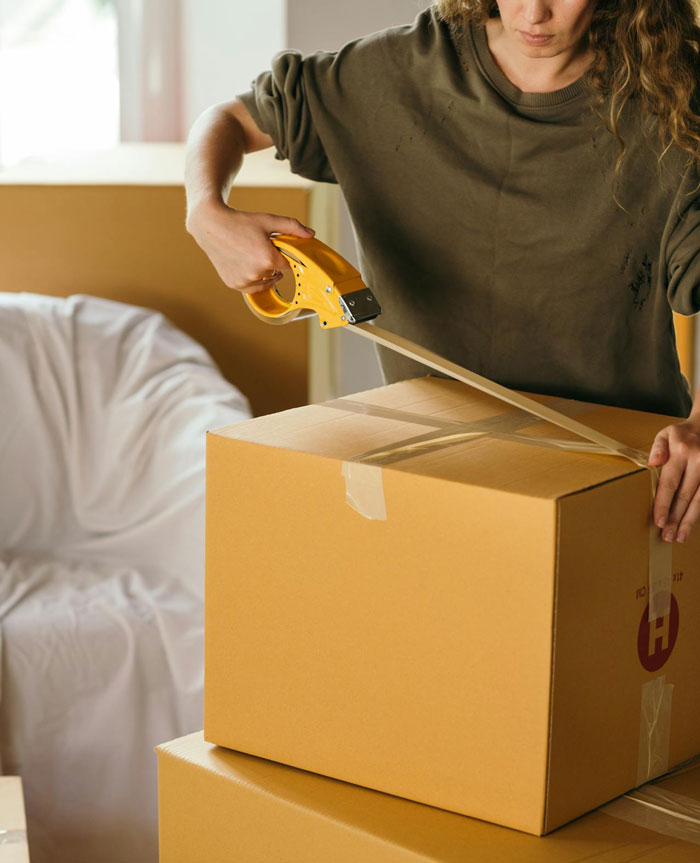 Woman packing boxes using tape dispenser, symbolizing husband left picking up the pieces after wife leaves toddlers behind to party.