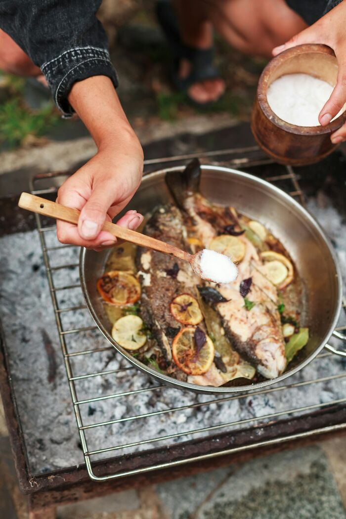 Hand sprinkling salt over grilled fish with lemon slices on an outdoor stove showcasing easy kitchen tricks.