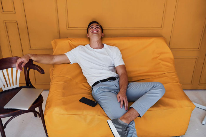Young man relaxing on yellow couch near chair with laptop and phone, representing conflict over care for autistic sibling.