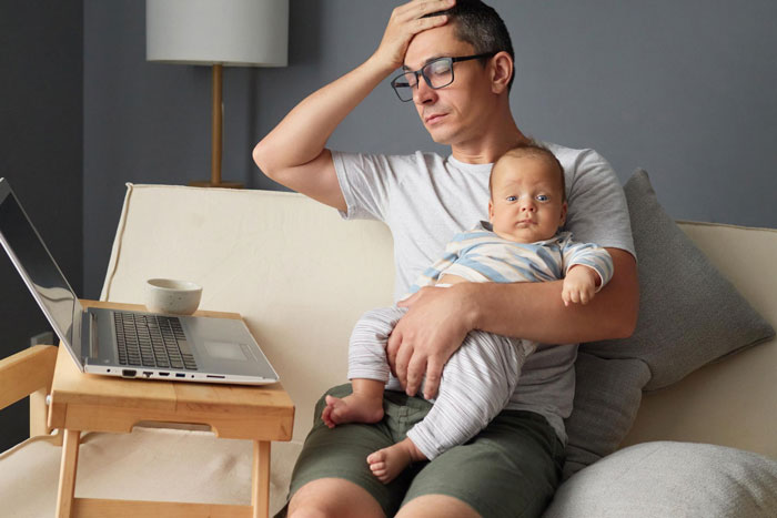 Stressed man holding baby on couch beside laptop, representing clash over care for autistic sibling in family dispute.