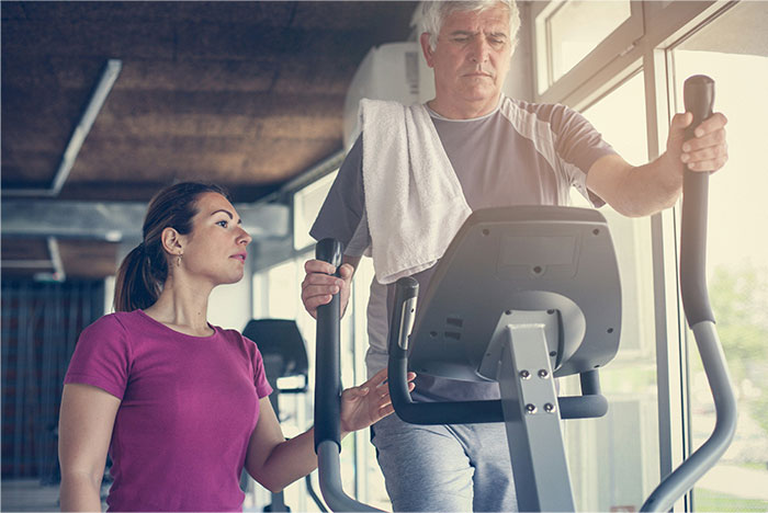 Man with towel on shoulder using elliptical machine while woman in purple shirt spots him in gym, illustrating fake illness exposure.