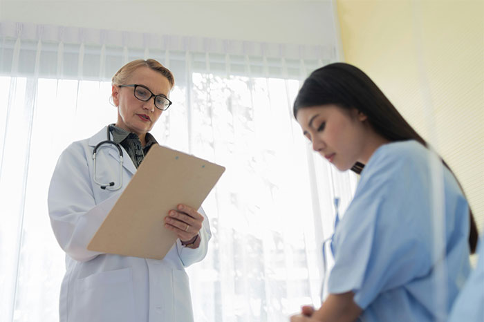 Doctor reviewing patient's chart in a bright medical office as patient in hospital gown looks concerned about illness.