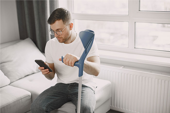 Man with crutch checking his phone while sitting on a couch, illustrating attempts to fake illnesses and getting exposed.