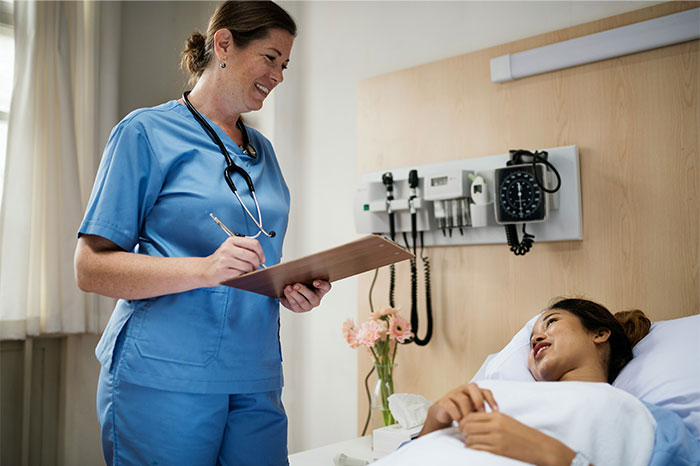 Nurse in blue scrubs smiling and taking notes while talking to a patient lying in a hospital bed, illustrating faked illnesses.