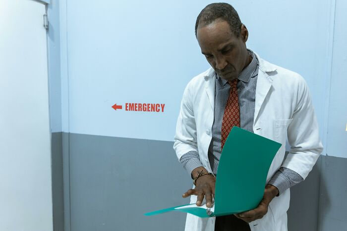 Doctor in white coat reviewing patient files near emergency sign, illustrating patients trying to fool doctors with ridiculous lies.