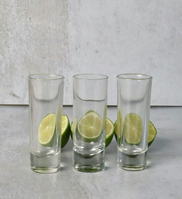 Three empty shot glasses in a row with lime wedges behind them on a light gray surface and background.