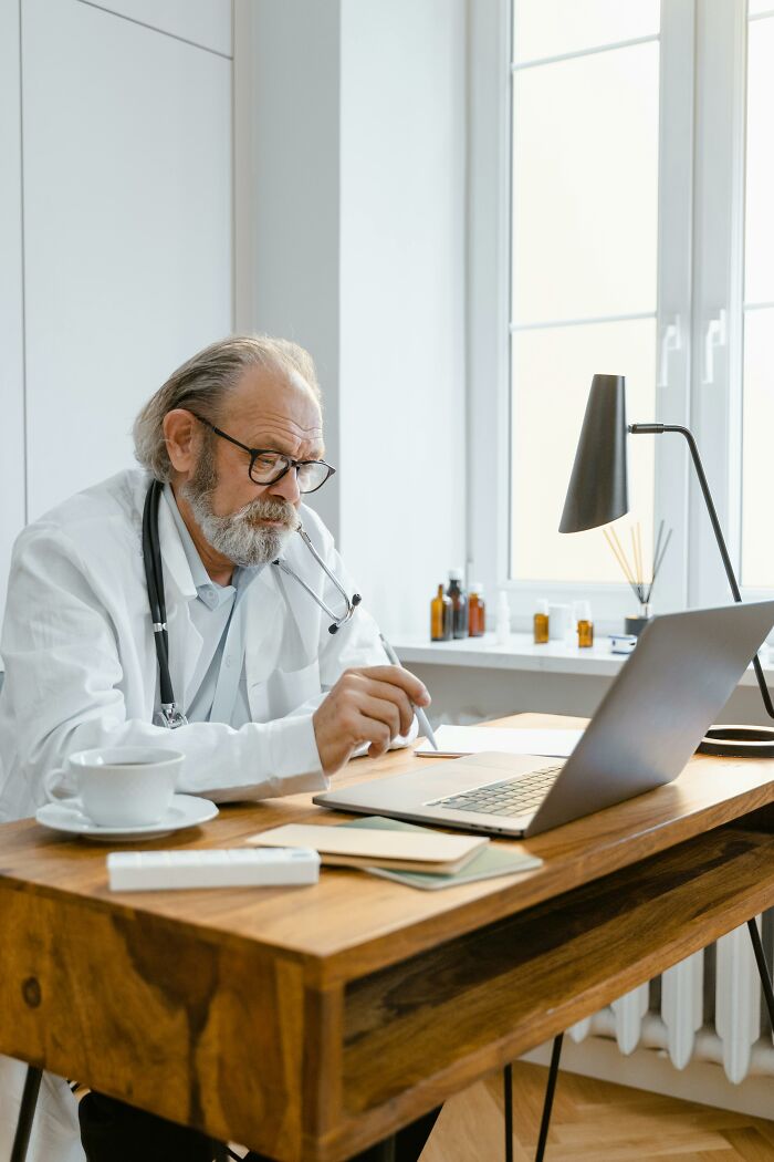 Older male doctor with glasses and stethoscope working on laptop in a bright office, reviewing patient cases and medical notes.