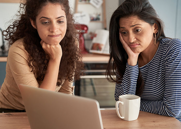 Two women looking puzzled and thoughtful while viewing a laptop, illustrating people living double lives scenarios.