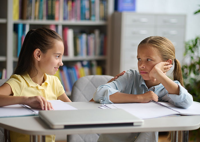 Two young girls at a table with notebooks, one comforting the other, illustrating moments of living double lives.