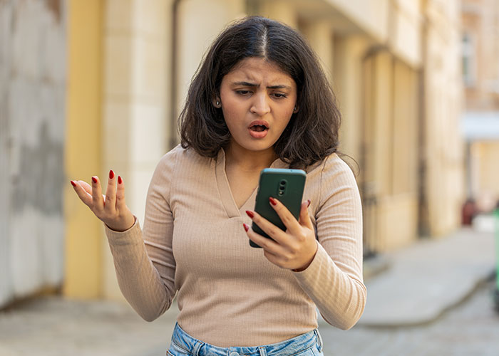 Young woman looking shocked at her phone, reacting to someone trying to live double lives and fool others outdoors.
