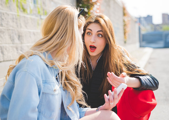 Two young women sit outdoors, one showing a phone to the other with a shocked expression about living double lives.